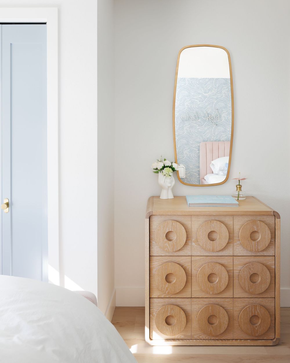 Light wood dresser with circular carved drawer fronts topped with a gold-framed mirror in a bright white bedroom.