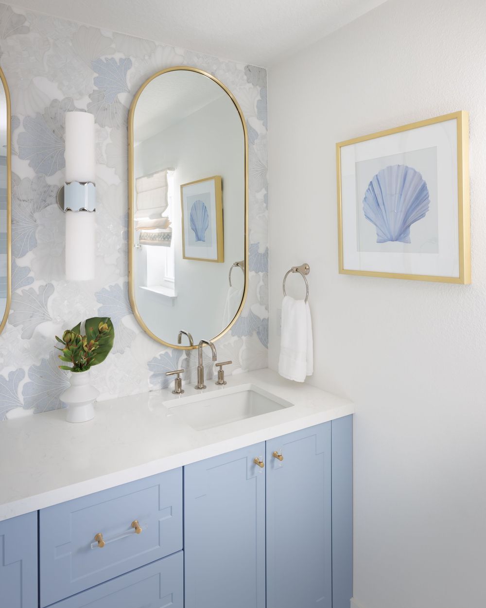 Blue vanity cabinet with white countertop, gold oval mirror, and seashell wallpaper in a coastal-style bathroom.