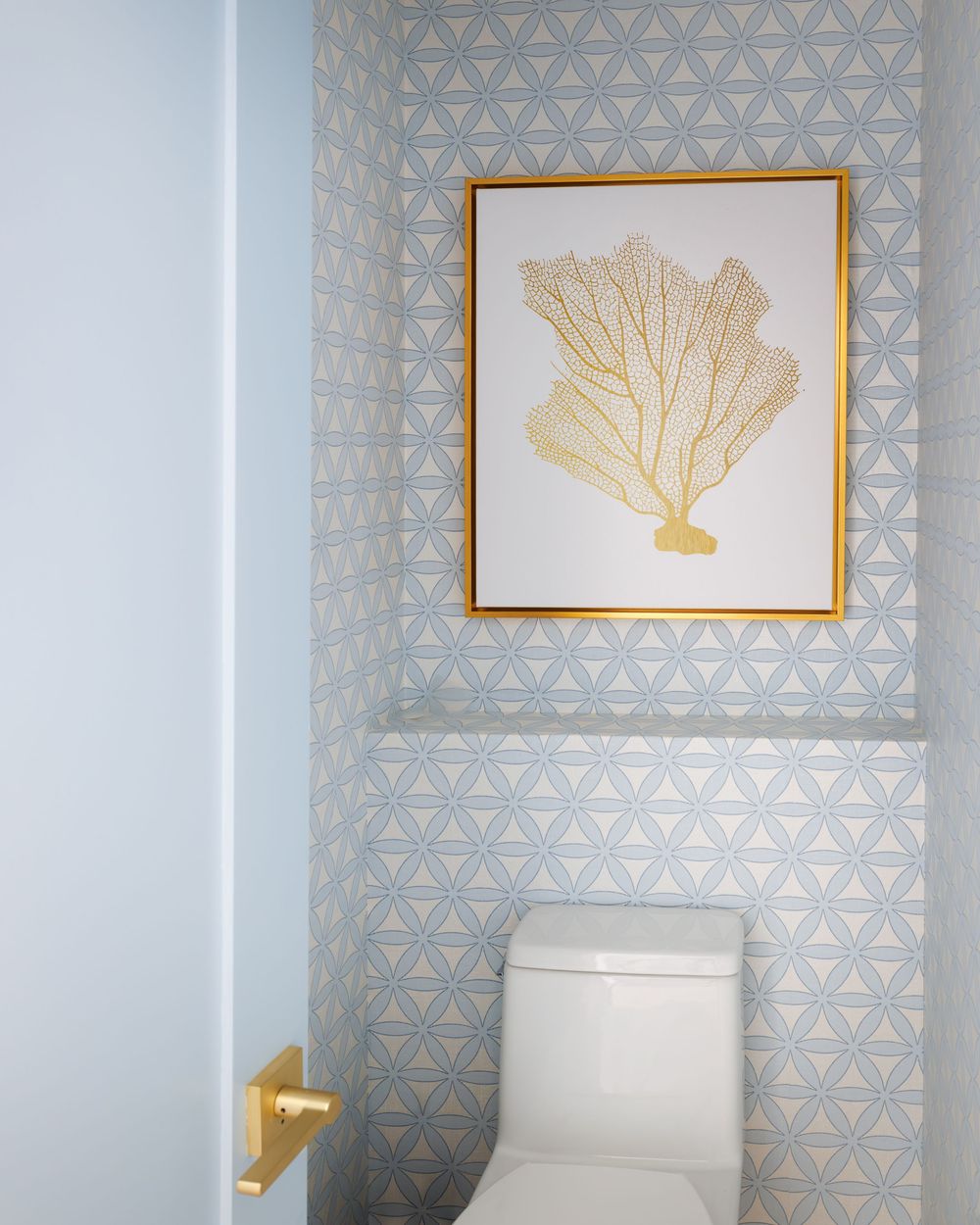 Light blue geometric wallpaper toilet nook with a gold-framed coral art print above the toilet.