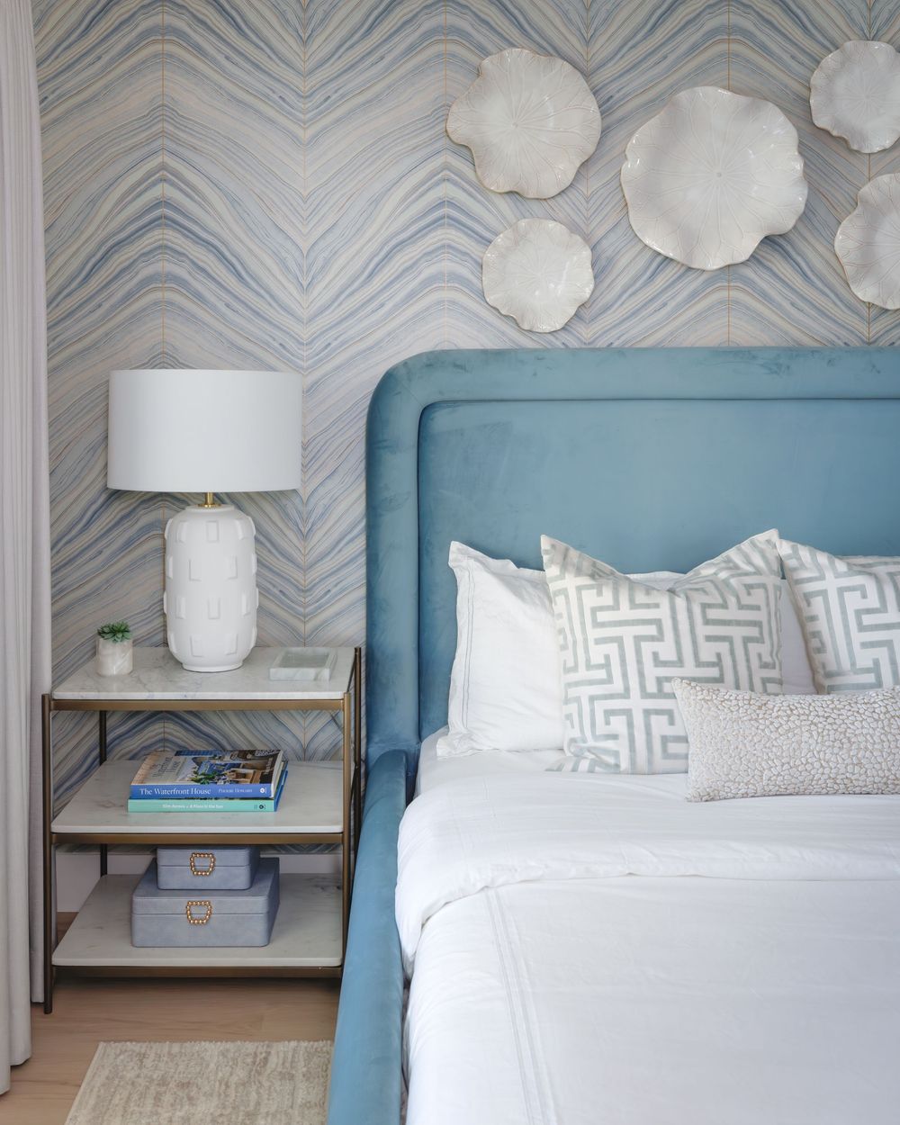 Blue upholstered headboard with geometric pillows against a blue wavy wallpaper accent wall in a serene bedroom.