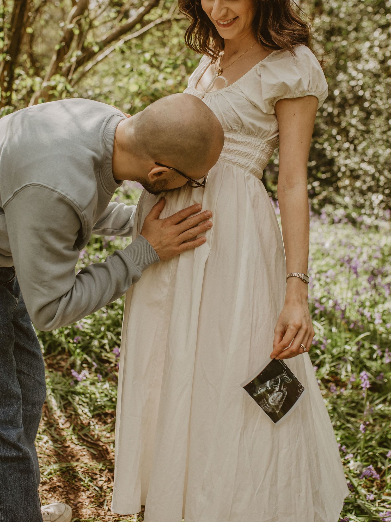 Man kisses pregnant woman's belly as she holds ultrasound photo in a flower-filled garden.