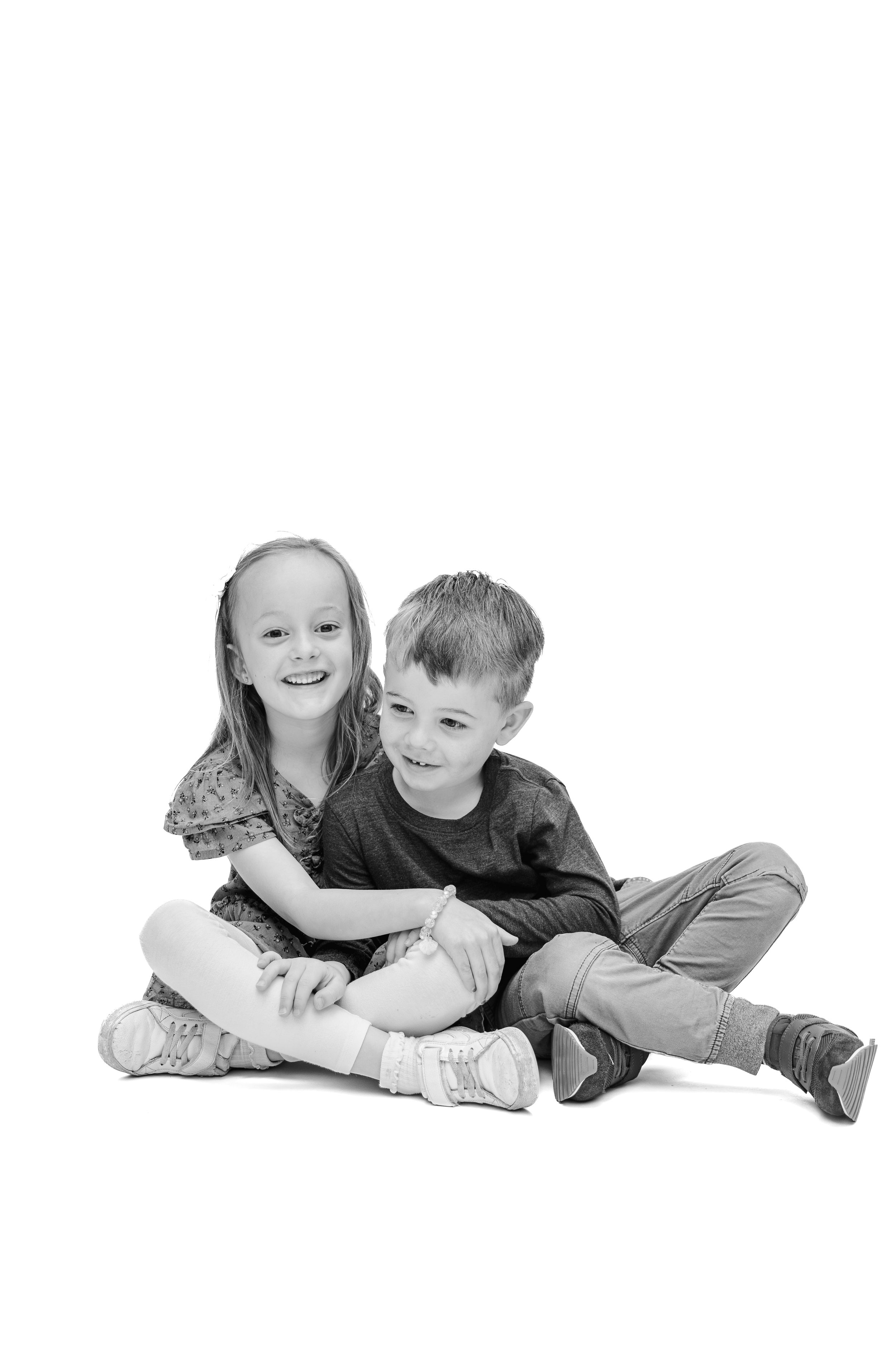Two happy children, a girl and boy, sit together on white background smiling in black and white photo.