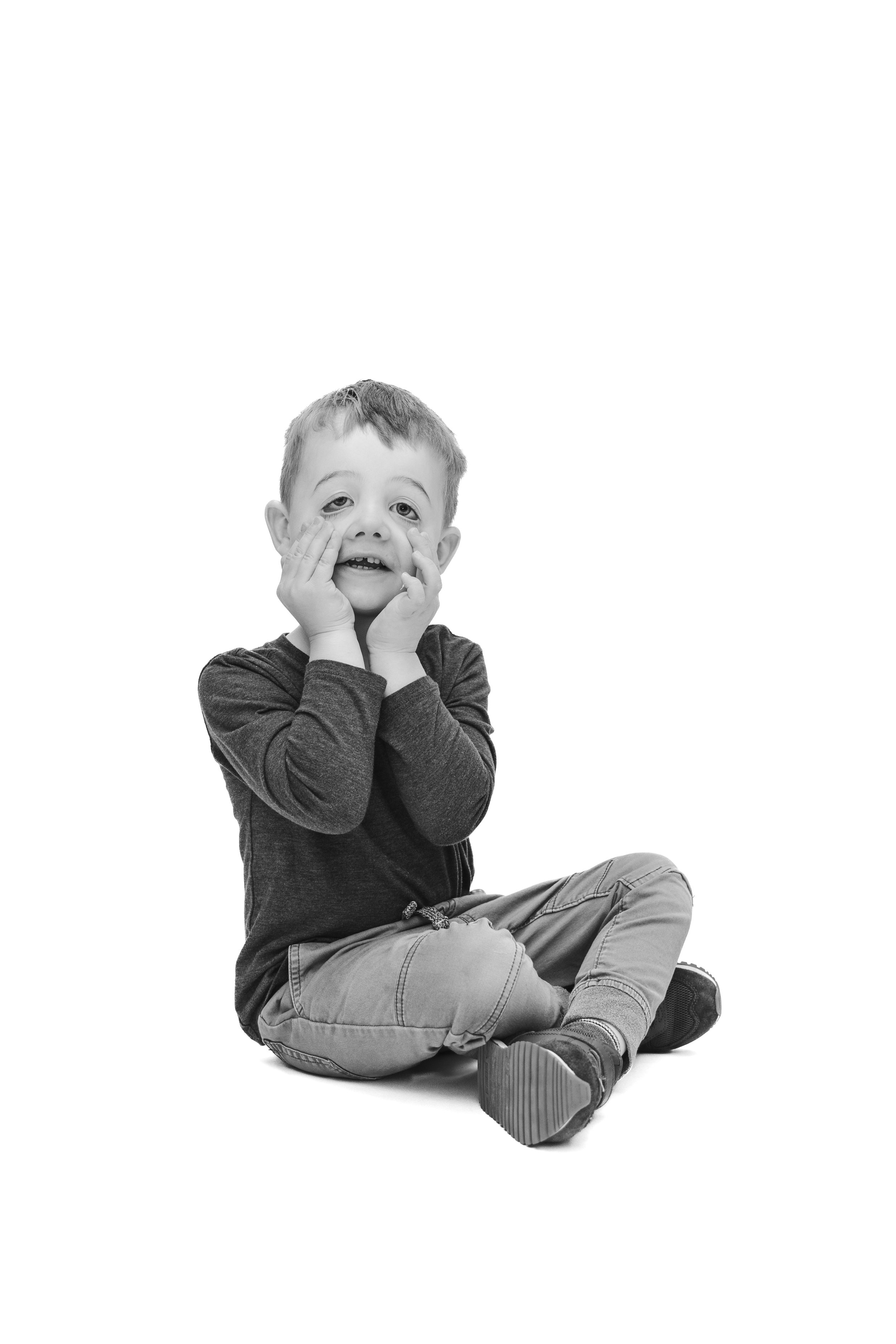 Black and white photo of a young boy sitting cross-legged with hands on cheeks, looking surprised.