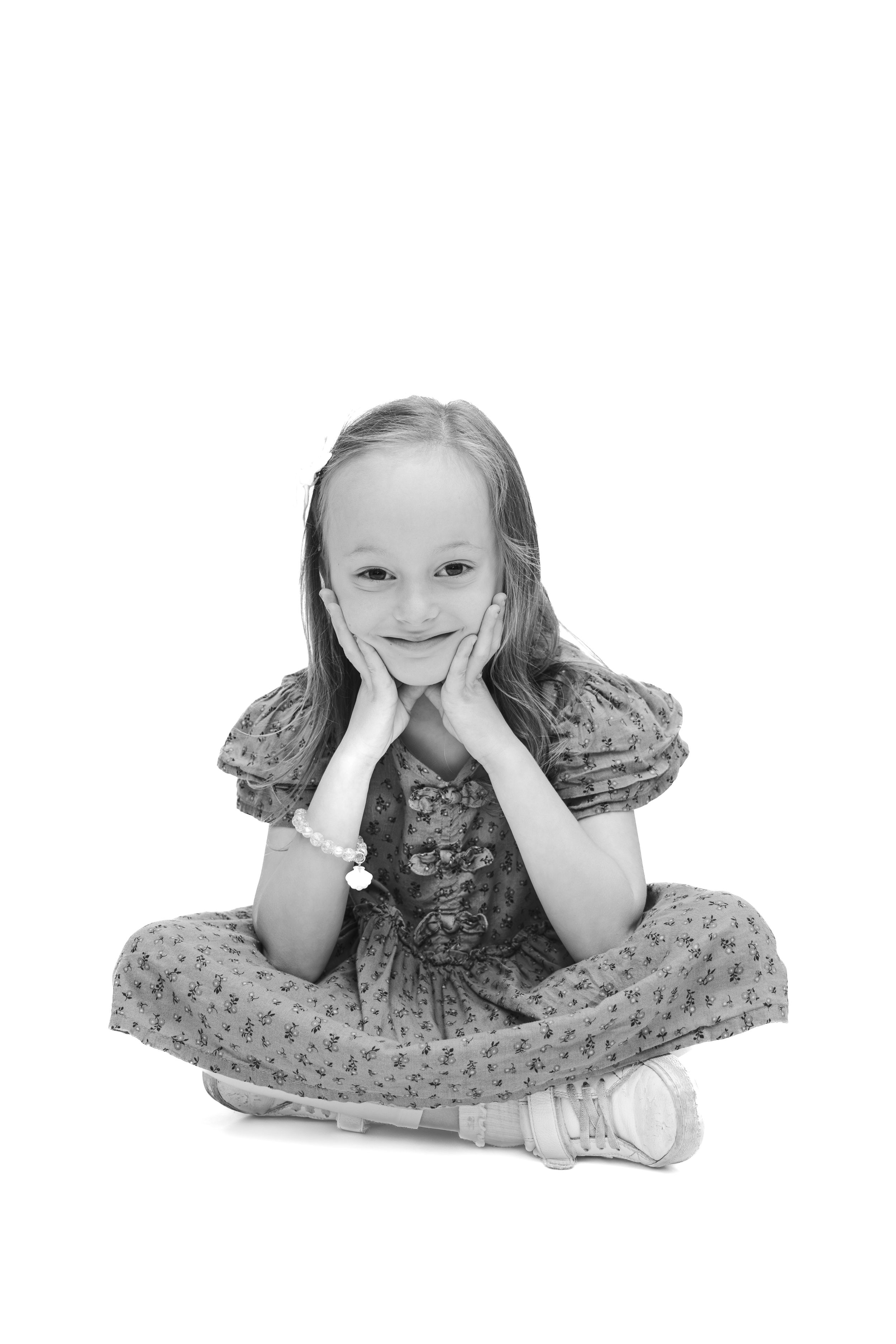 Black and white photo of a smiling young girl sitting cross-legged with hands on cheeks, wearing a floral dress.