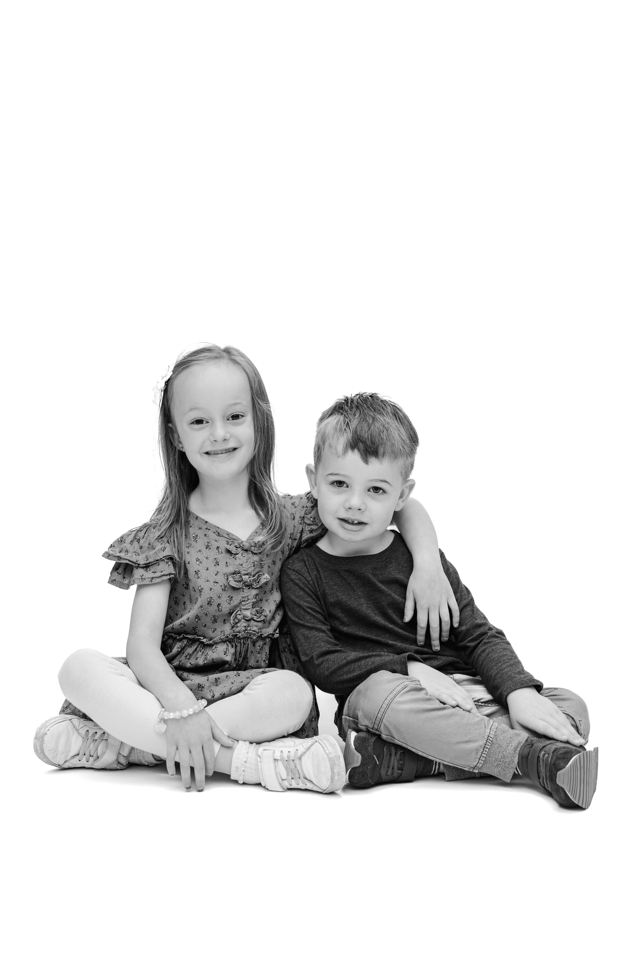 Black and white photo of two young siblings sitting together on white background, smiling at camera.