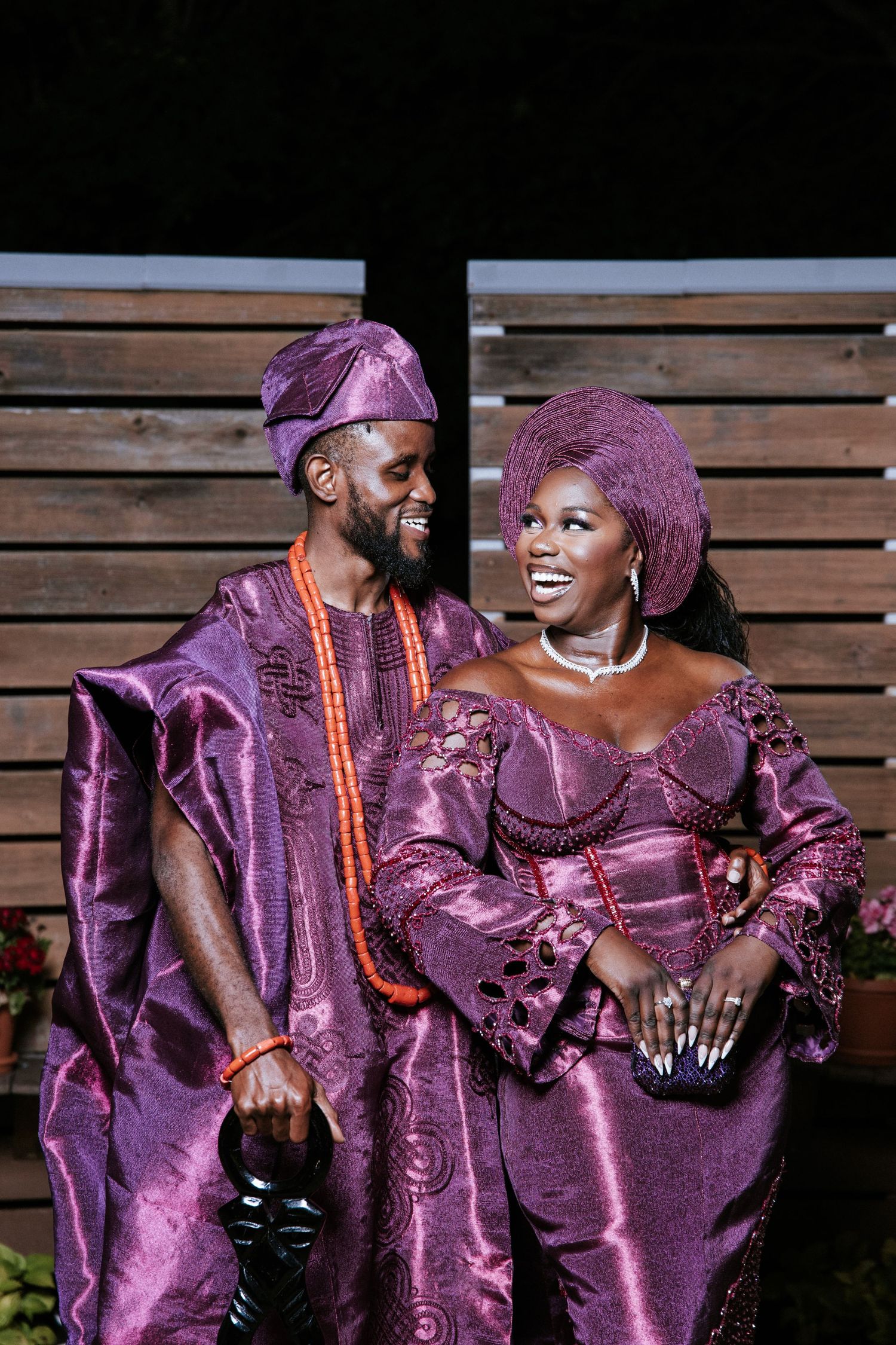 A joyful Black couple in vibrant African attire smile at each other under a white pergola outdoors.