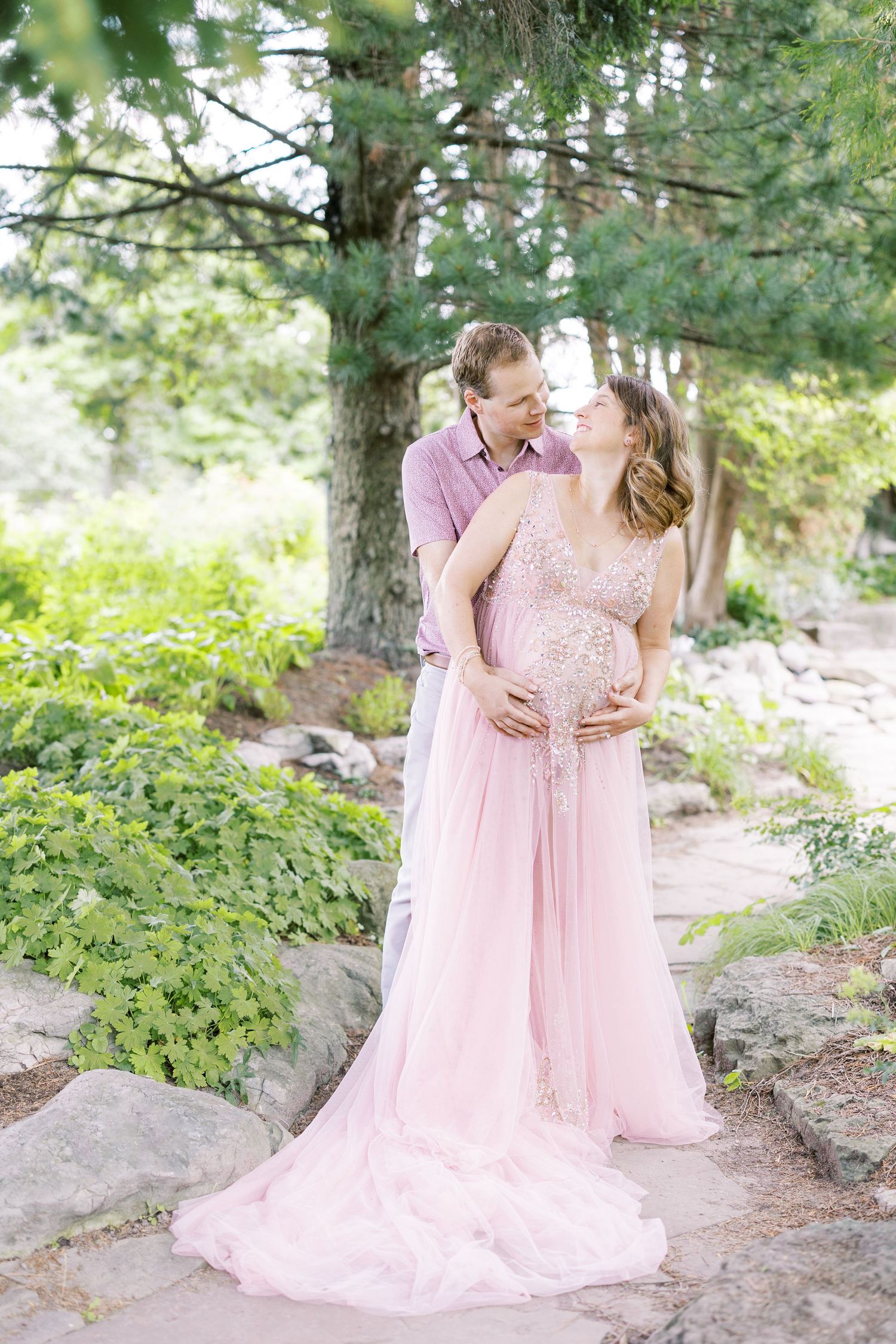 Couple in a garden, pregnant woman in pink gown, partner embracing her bump, surrounded by lush greenery.