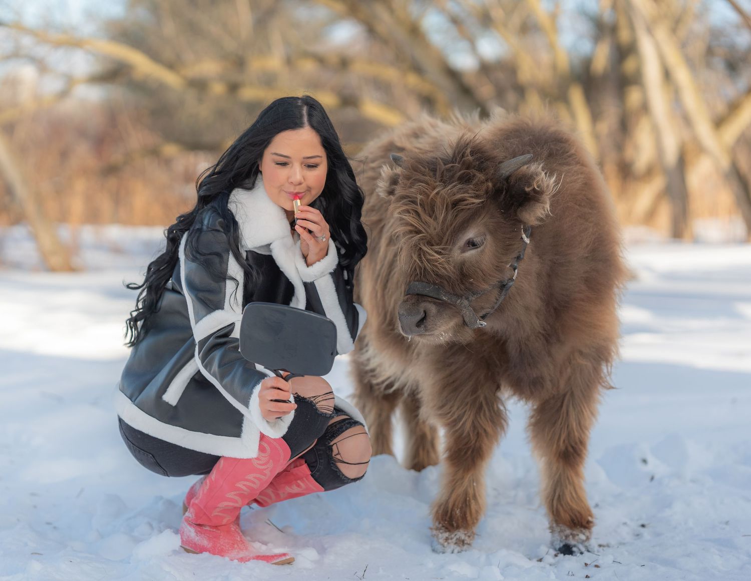 Woman in winter coat crouching beside a fluffy baby bison in a snowy landscape.