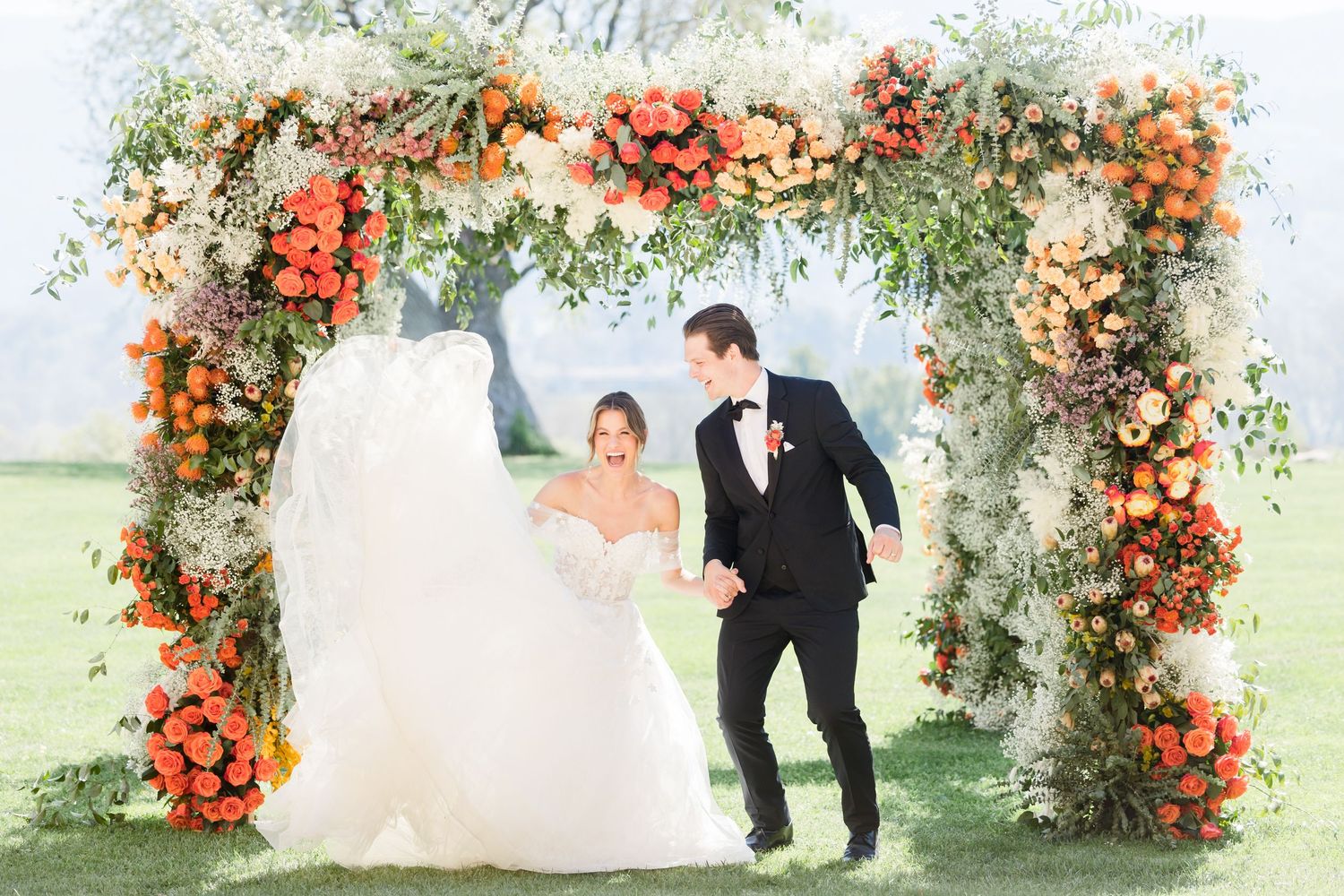 Bride and groom walking under a floral arch adorned with orange and white roses at an outdoor wedding ceremony.