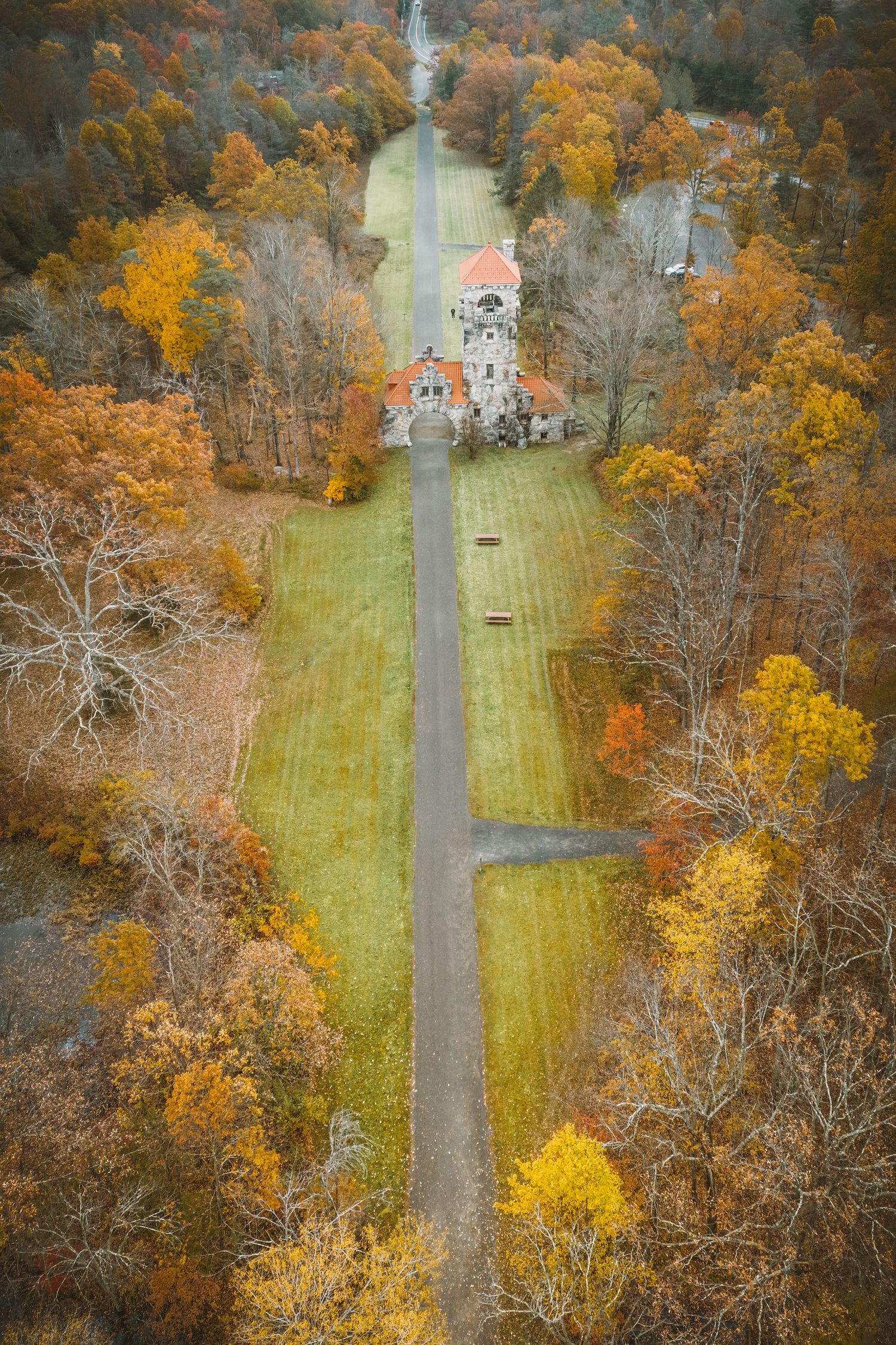 Aerial autumn view of a historic stone castle surrounded by colorful fall foliage along a long straight pathway.