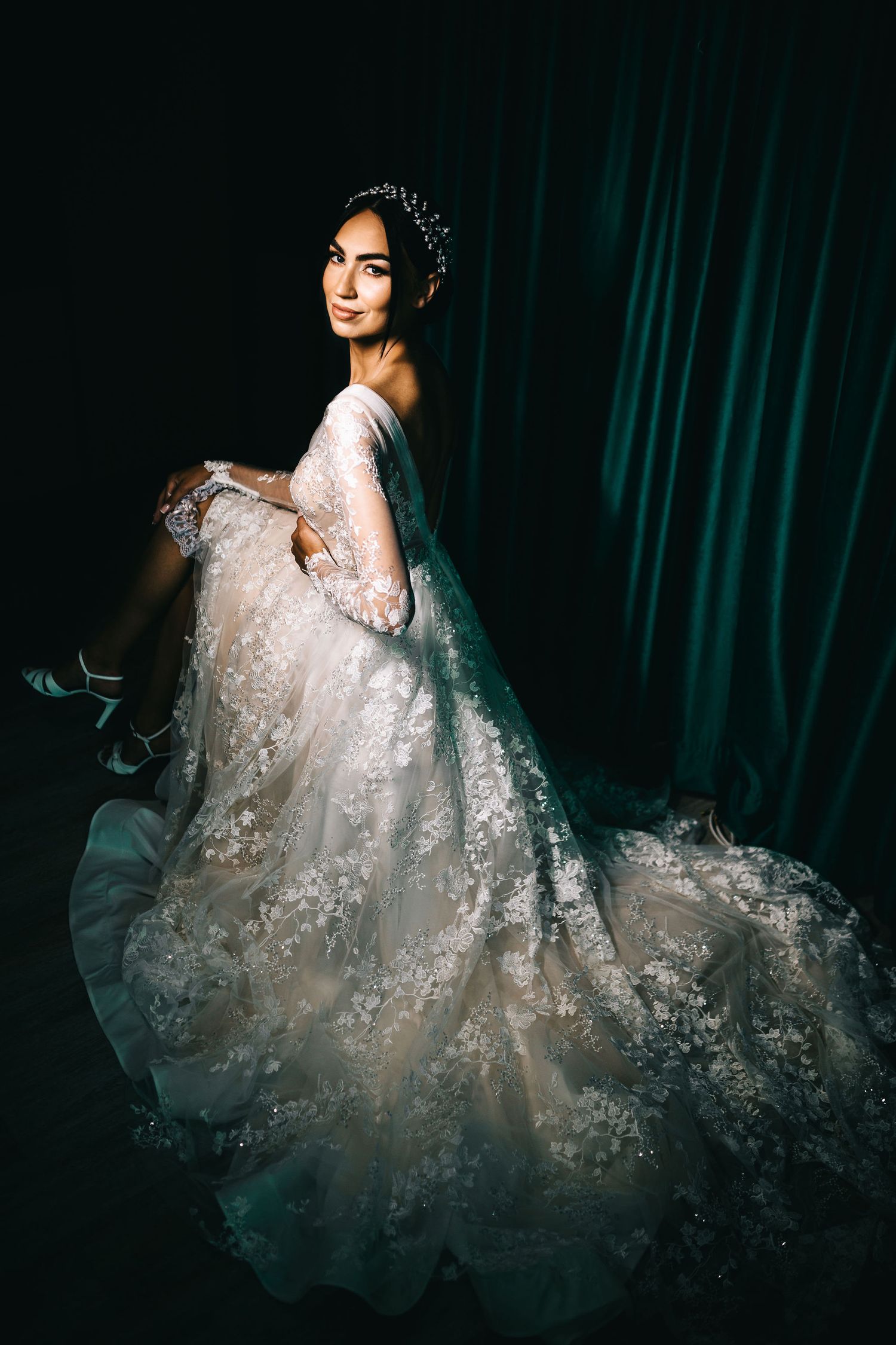 Elegant bride in lace wedding gown with crown sitting against dark teal curtain backdrop.