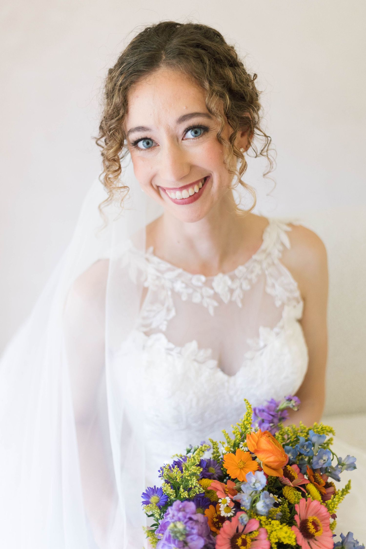 Bride in white lace gown and veil holds colorful bouquet of orange, purple, and blue flowers near a window.