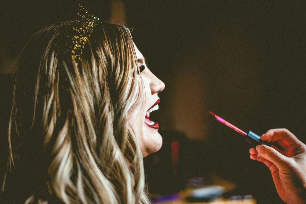 Woman with gold crown getting lipstick applied by makeup artist, mouth open, wavy blonde hair.