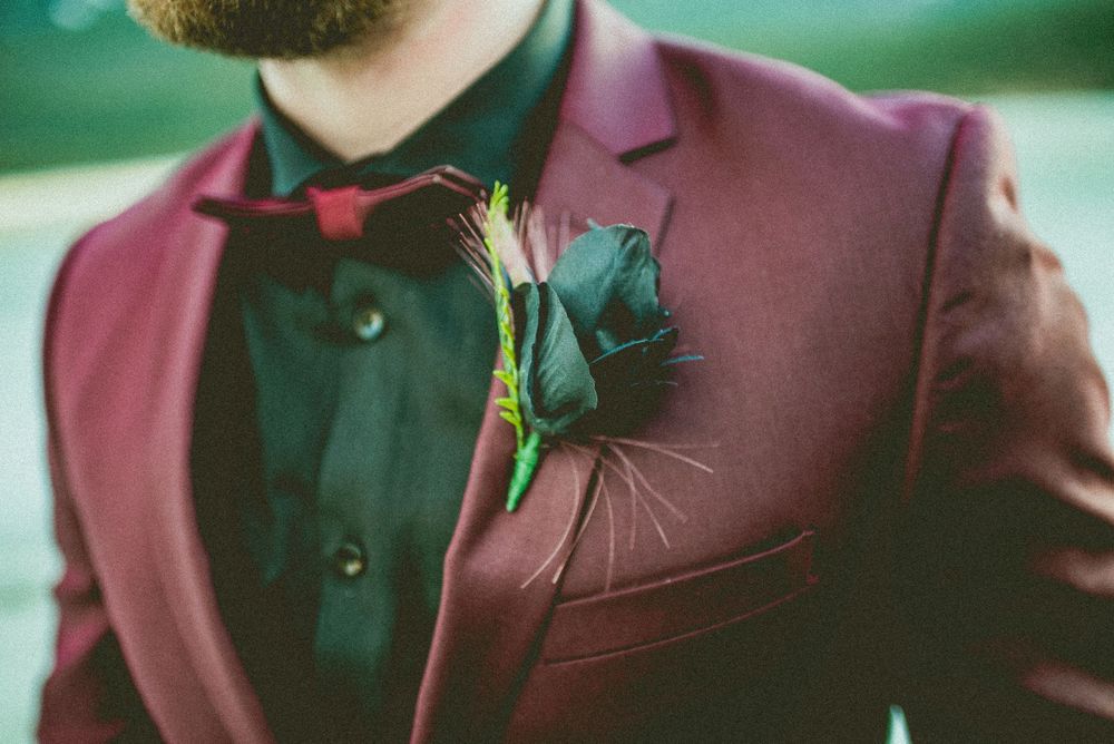 Man in burgundy suit with black rose boutonniere and red bow tie, elegant wedding groom fashion detail.
