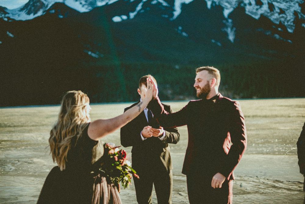 Couple high-fiving during outdoor winter wedding ceremony with snowy mountains in background.
