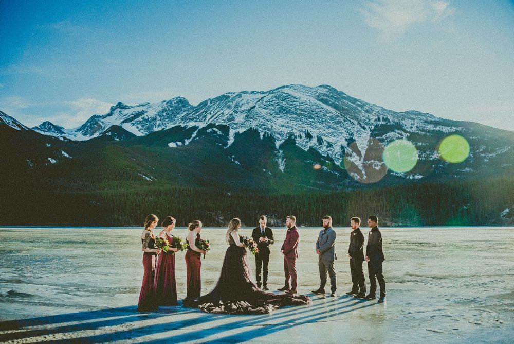 Wedding party poses on frozen lake with snow-capped mountains, bridesmaids in burgundy gowns and groomsmen in suits.