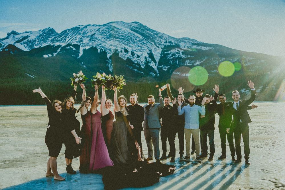 Wedding party celebrating outdoors with snow-capped mountains backdrop, bridesmaids in burgundy dresses raising bouquets.