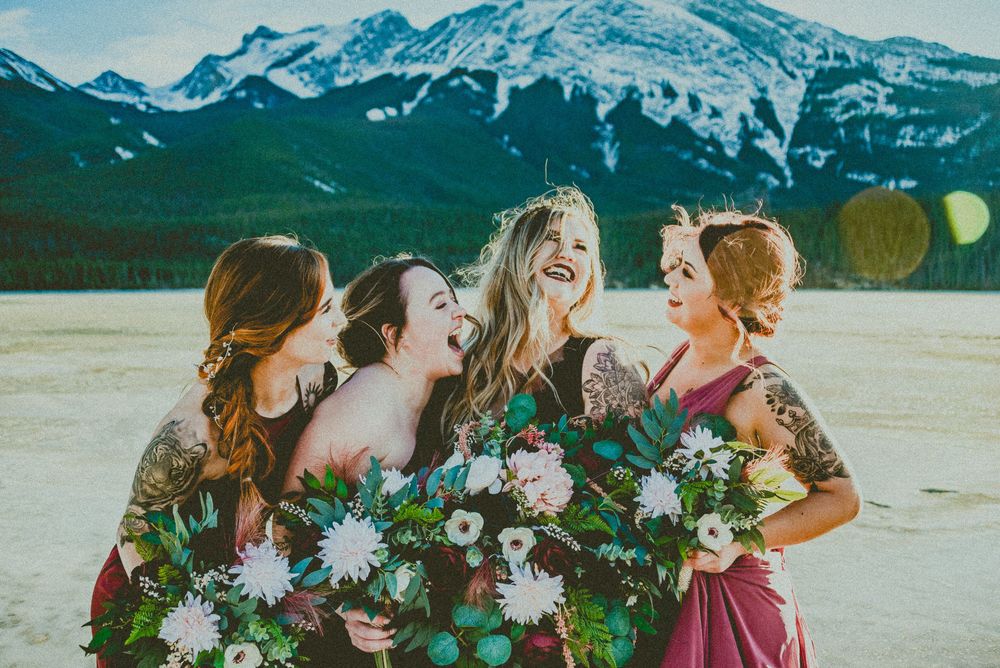 Four bridesmaids in burgundy dresses laughing together holding bouquets with snow-capped mountains in the background.