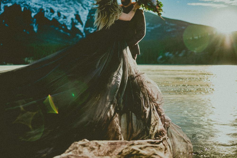 Woman in flowing black gown standing by a mountain lake at sunset with golden light.