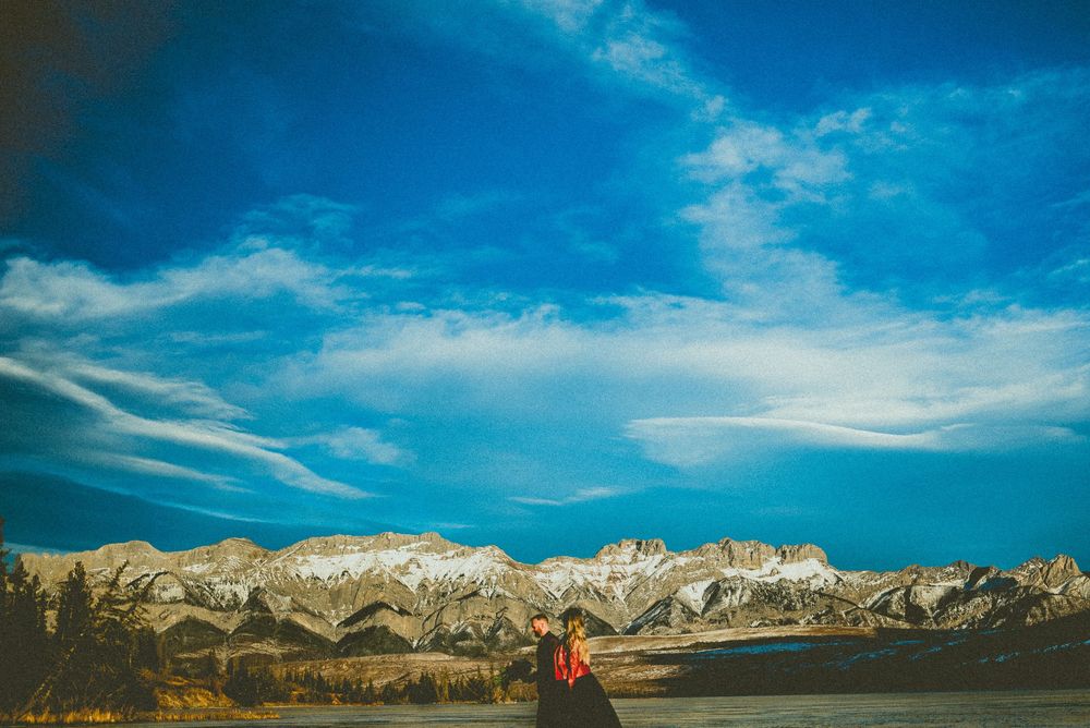 Couple embracing against a dramatic blue sky with snow-dusted mountain range in the distance.