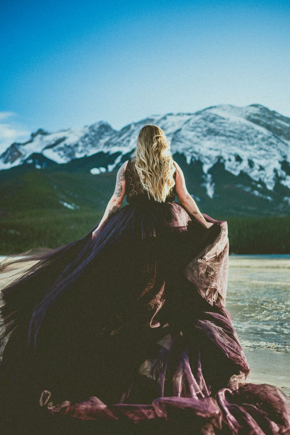 Bride in flowing dark purple gown standing on rocks facing a snow-capped mountain lake.