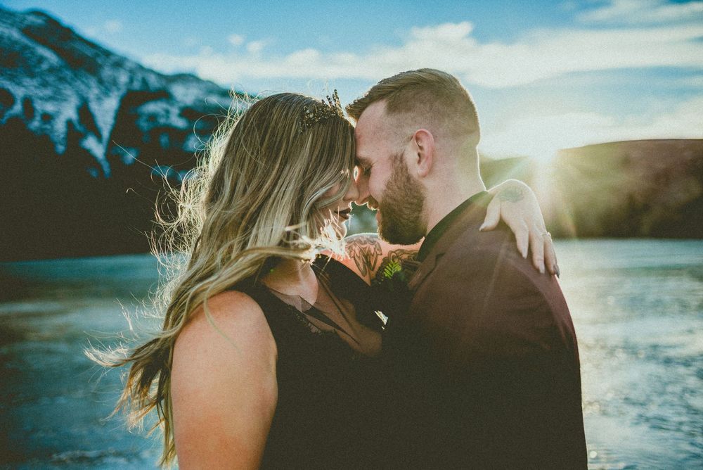 Romantic couple sharing an intimate moment by a mountain lake at sunset, dressed in elegant wedding attire.