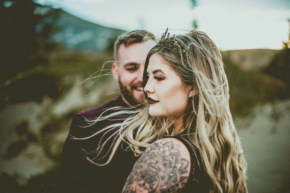 Tattooed bride in black crown embraces bearded groom in moody outdoor wedding portrait.