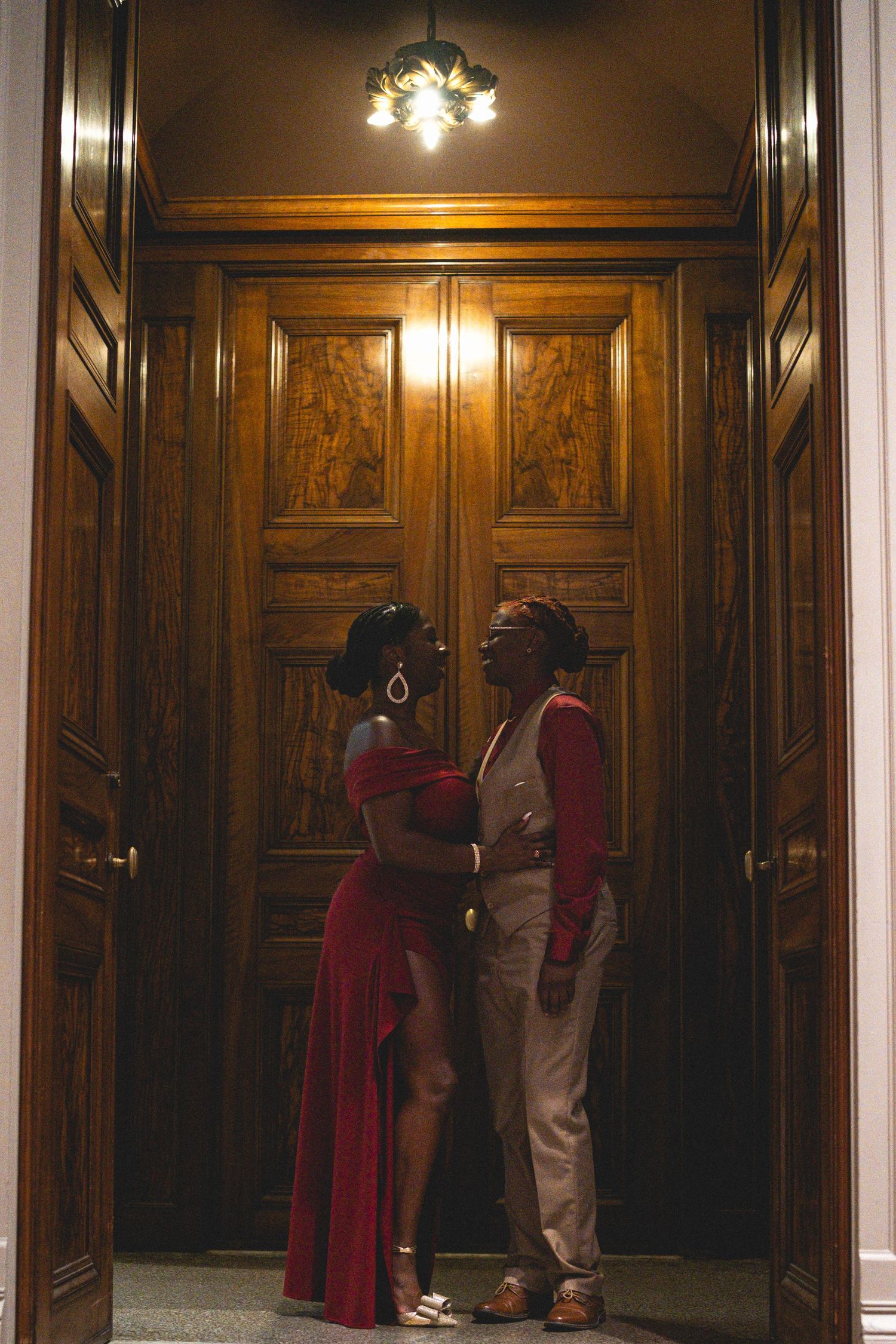 Couple in formal attire sharing a romantic moment in front of elegant wooden doors under warm chandelier light.