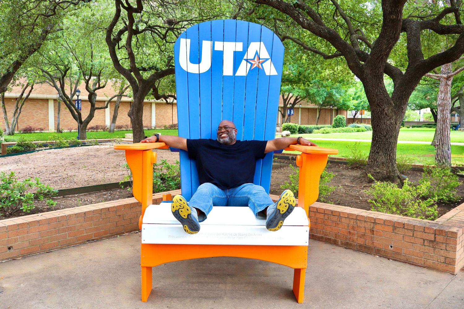 A man sits in a large blue and orange UTA chair outdoors on a sunny day.