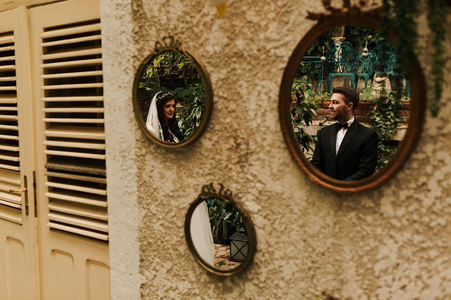 Bride and groom stand by window, groom holding white rose bouquet, bride wearing veil and floral crown.