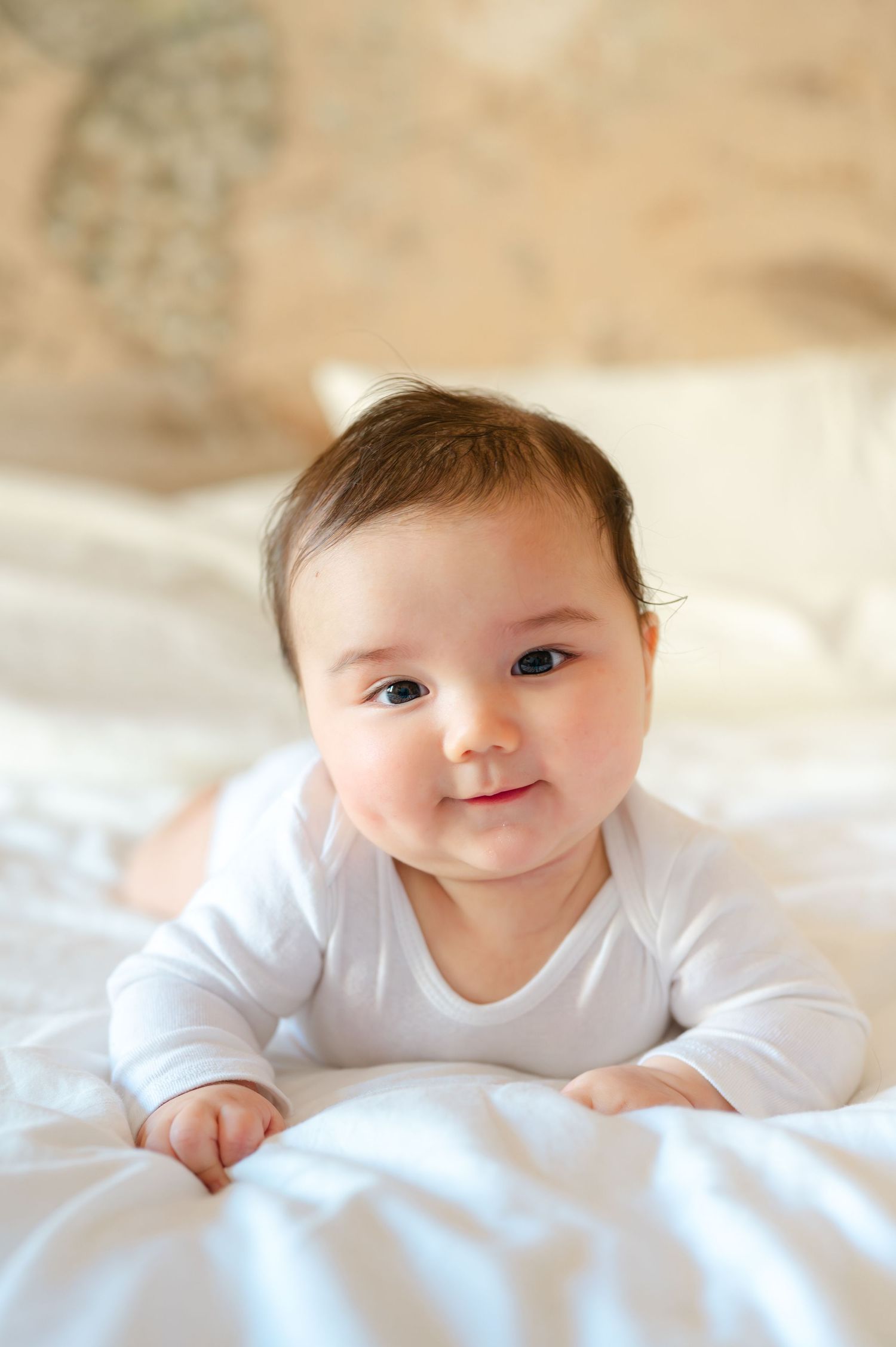 Adorable baby in white onesie doing tummy time on white bed, smiling at camera with bright eyes.