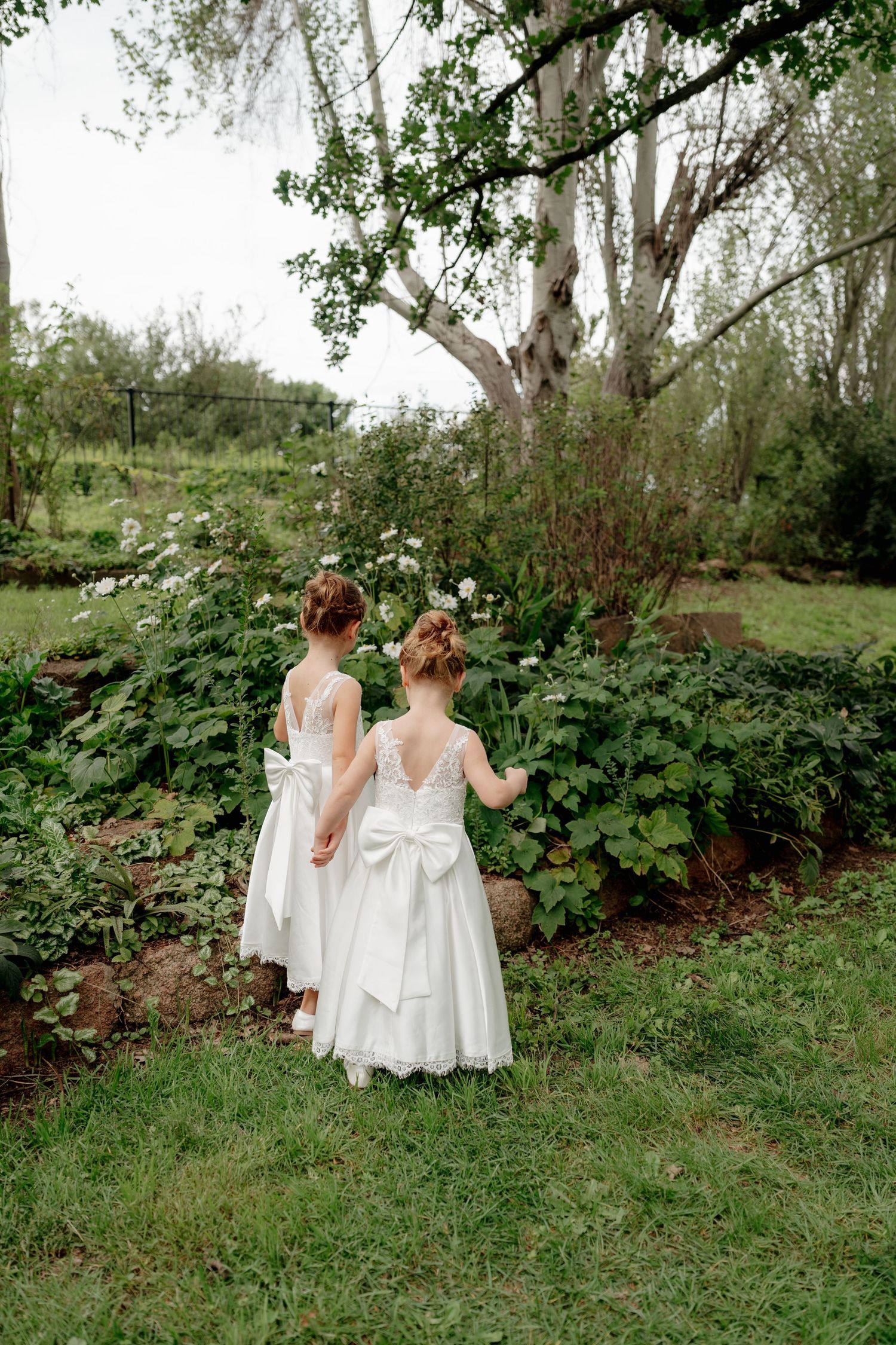 Two flower girls in white dresses with bow backs hold hands in a lush garden setting.