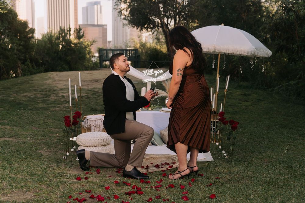 Man proposes to woman on one knee outdoors with rose petals, candles, and city skyline backdrop.