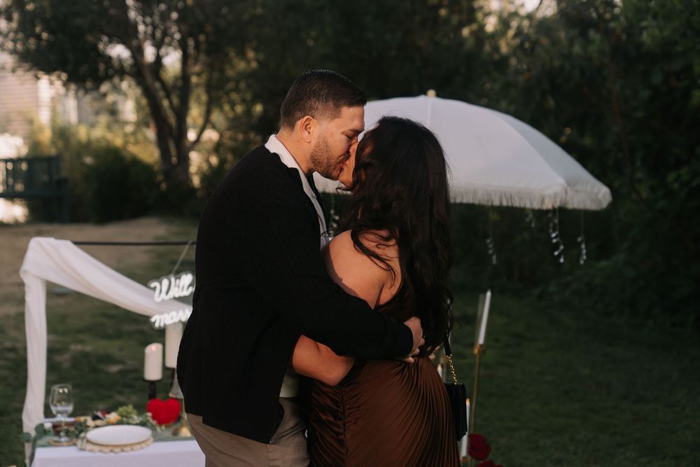 Couple sharing a romantic kiss outdoors at an elegant garden setup with white decor and roses at golden hour.