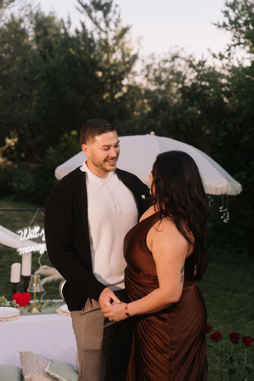 A couple holds hands and smiles at each other during an outdoor romantic dinner setup at golden hour.