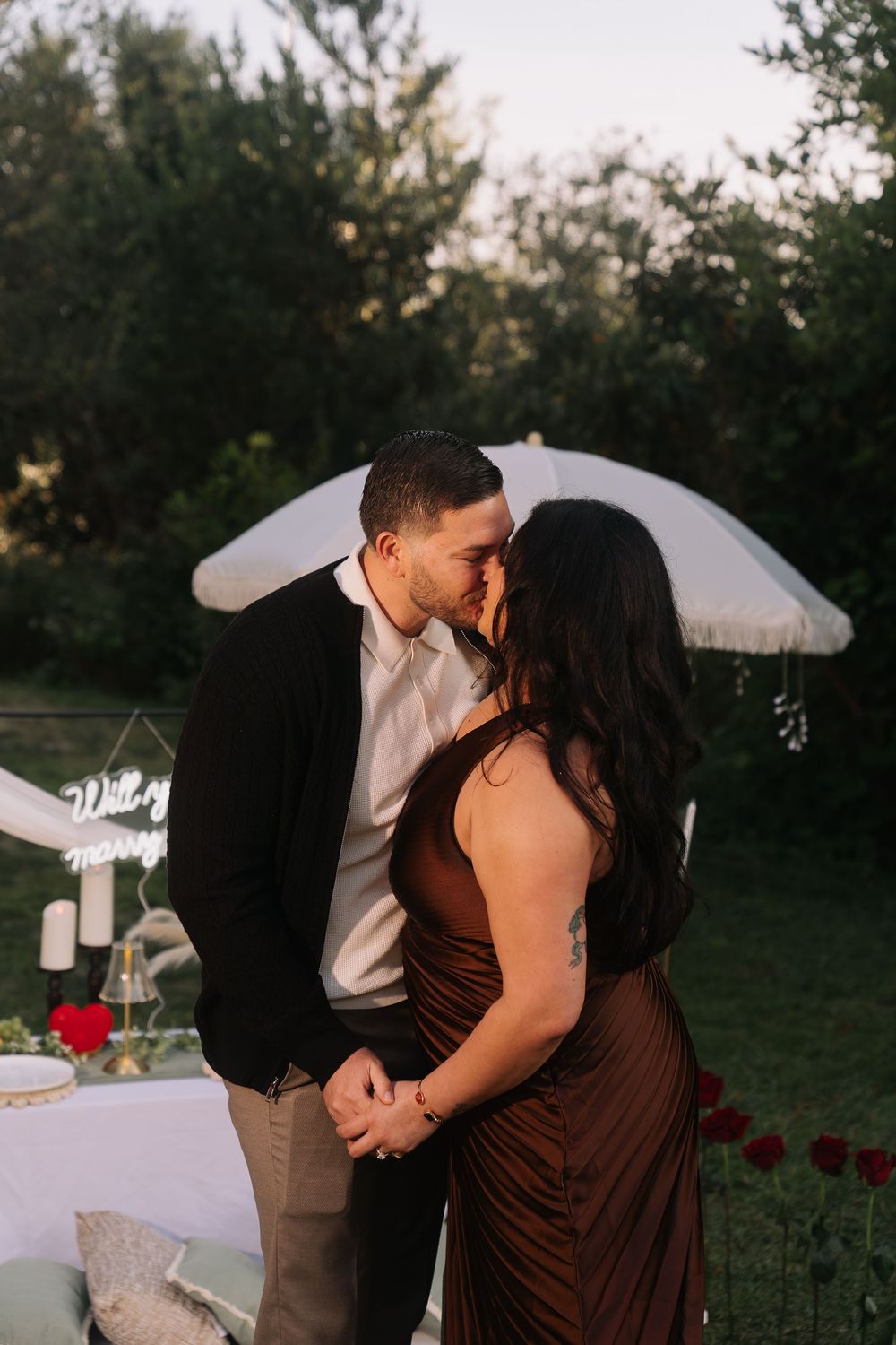 Couple sharing a romantic kiss during an outdoor proposal setup with a Will You Marry Me sign and candles at sunset.