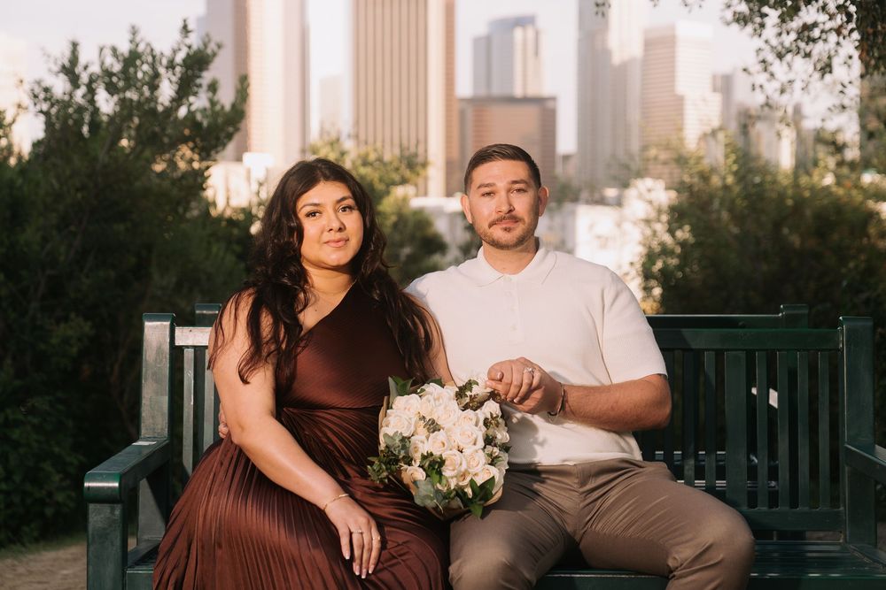 Couple sitting on a bench with city skyline backdrop, woman in brown dress holding white bouquet, man in white shirt.