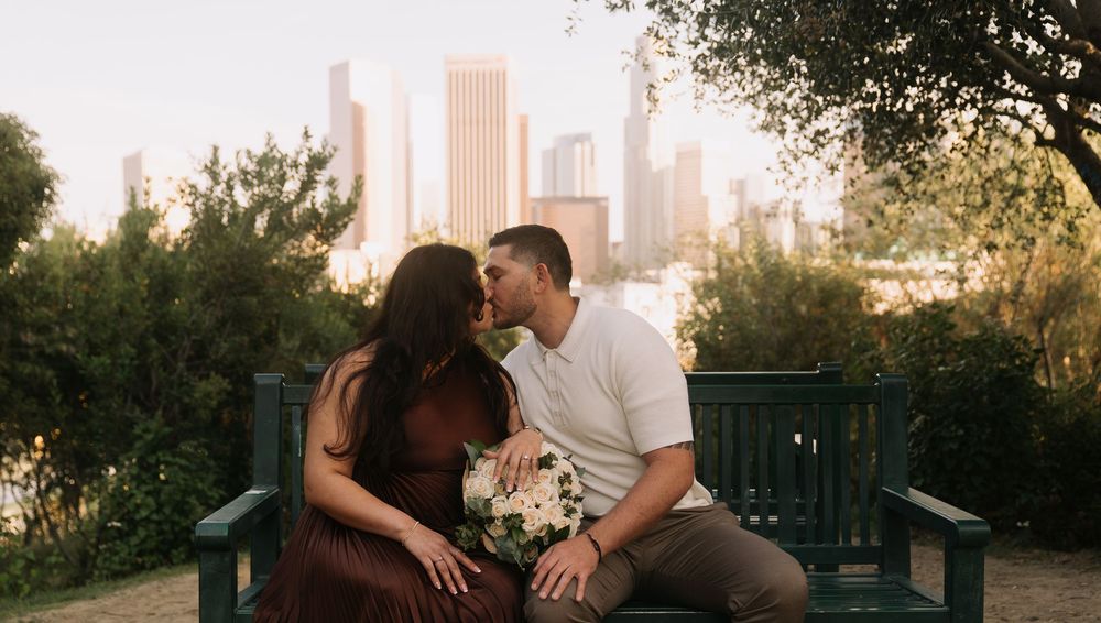 Couple sharing a romantic kiss on a park bench with a city skyline and lush green trees in the background.
