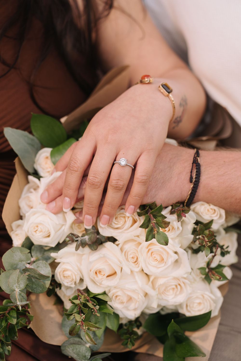 Couple's hands resting on white rose bouquet, woman wearing diamond engagement ring, man with bracelets.