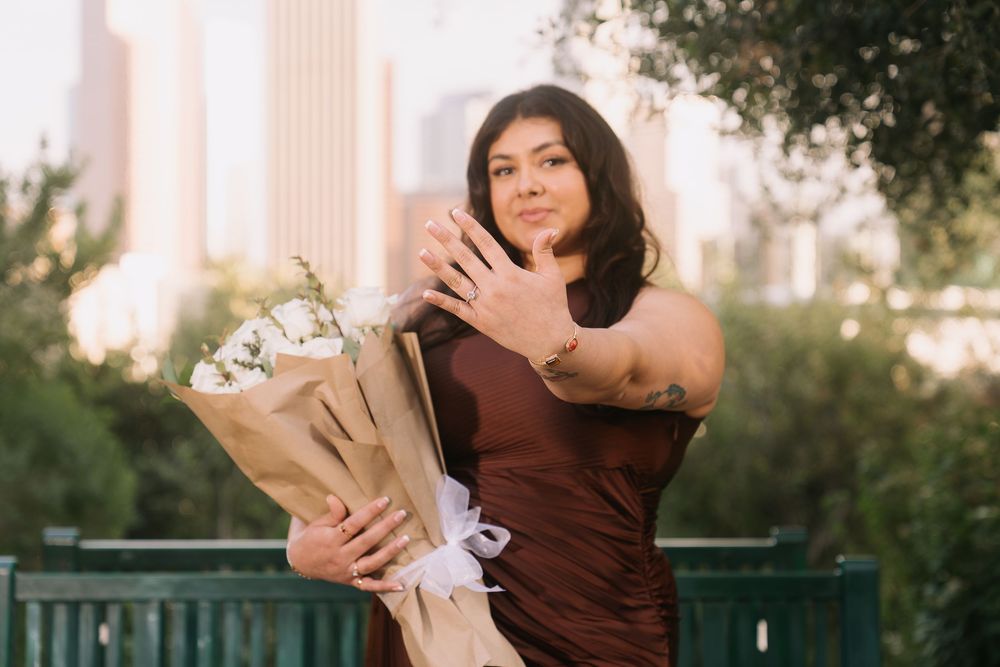 Woman in burgundy dress holding white flower bouquet, showing ring outdoors with city skyline background.