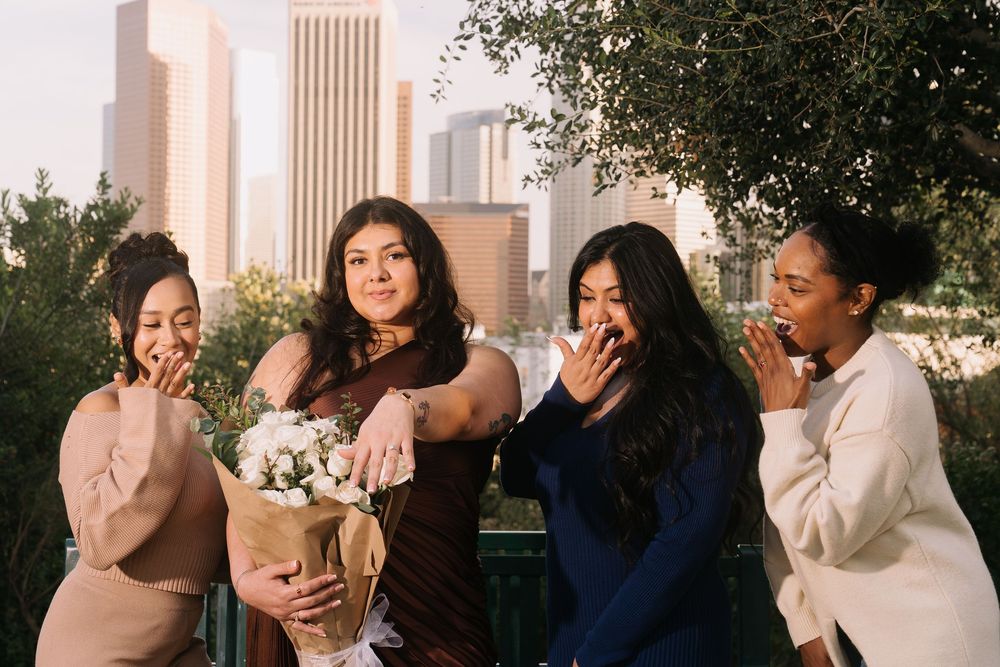 Four women pose outdoors with city skyline, one holding white flowers, all reacting with surprise and delight.