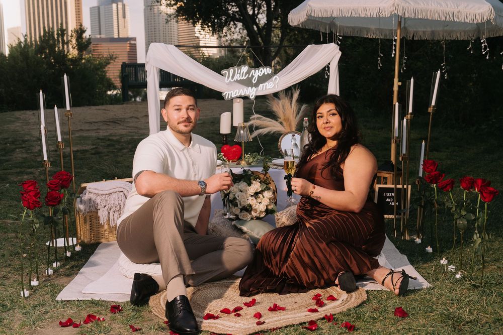 Couple sitting on a romantic outdoor picnic setup with red roses, candles, and a will you marry me sign at dusk.