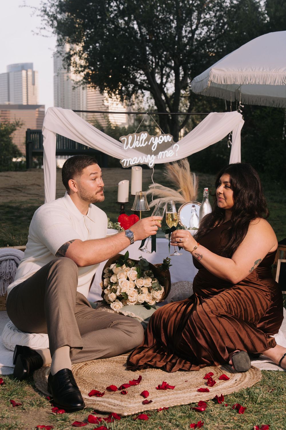 Couple toasting champagne at romantic outdoor proposal setup with Will You Marry Me sign, rose petals, and white flowers.