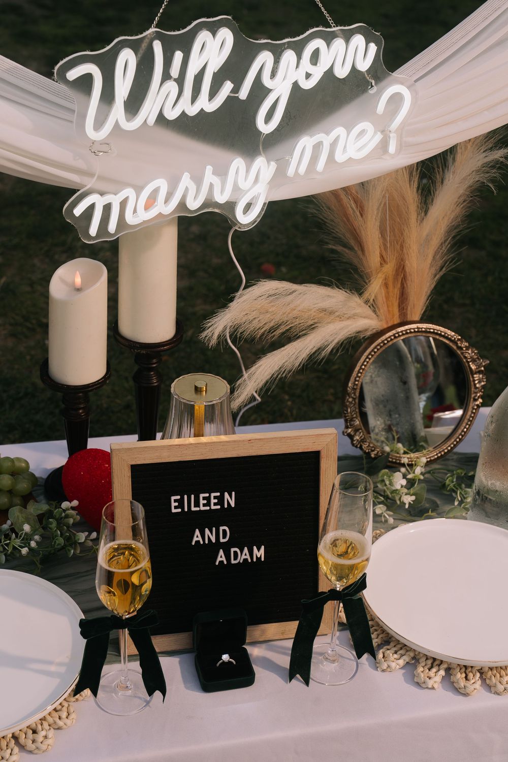 Romantic proposal setup with neon Will You Marry Me sign, champagne glasses, and letter board reading Eileen and Adam.