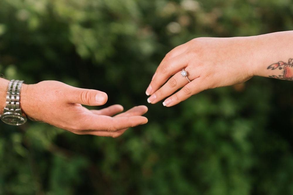 Two hands reaching toward each other, one wearing a diamond ring and one with bracelets, against green foliage.