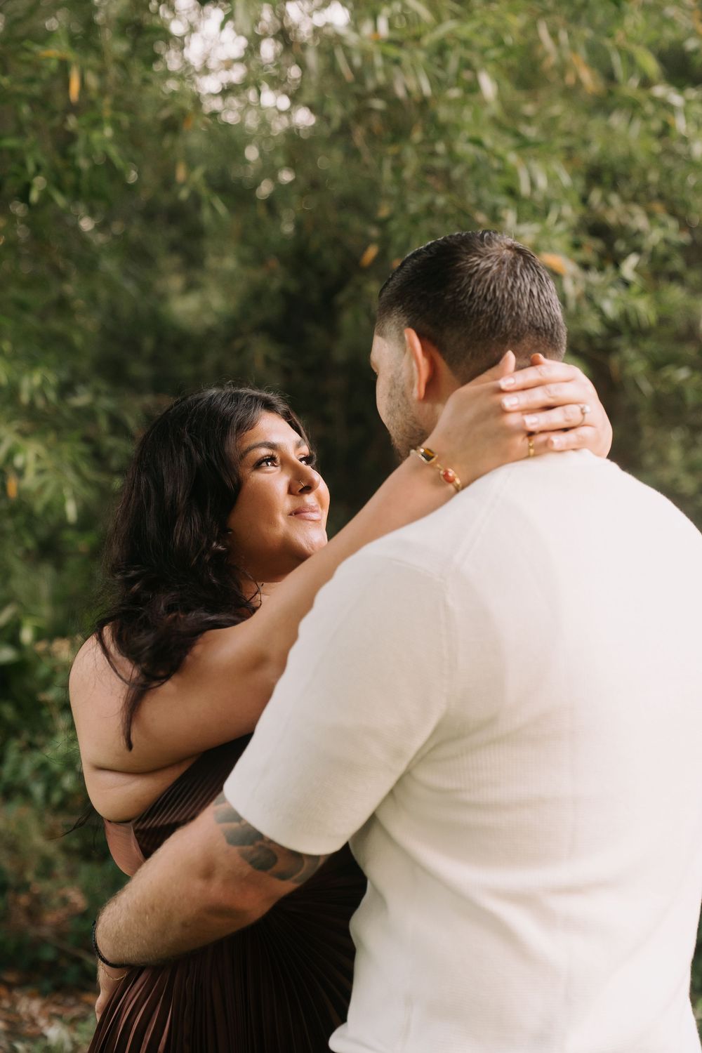 A couple embraces outdoors, woman in brown dress smiling at man in white shirt, surrounded by lush green foliage.