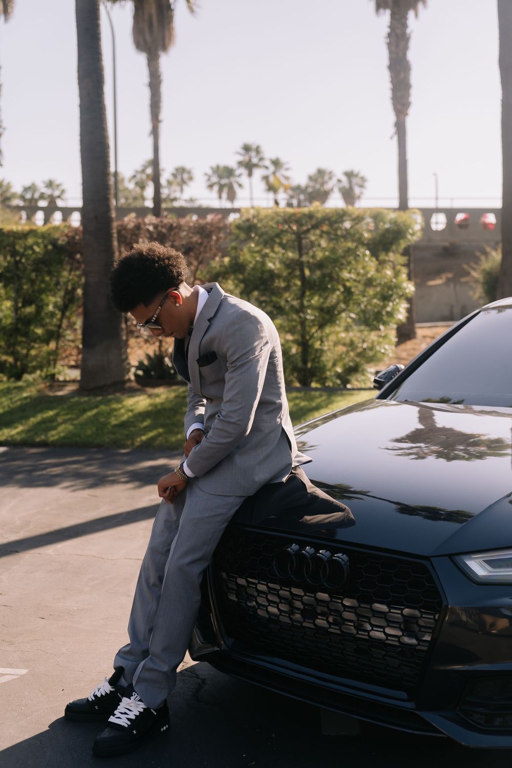 Young man in gray suit leaning on black Audi car surrounded by palm trees.