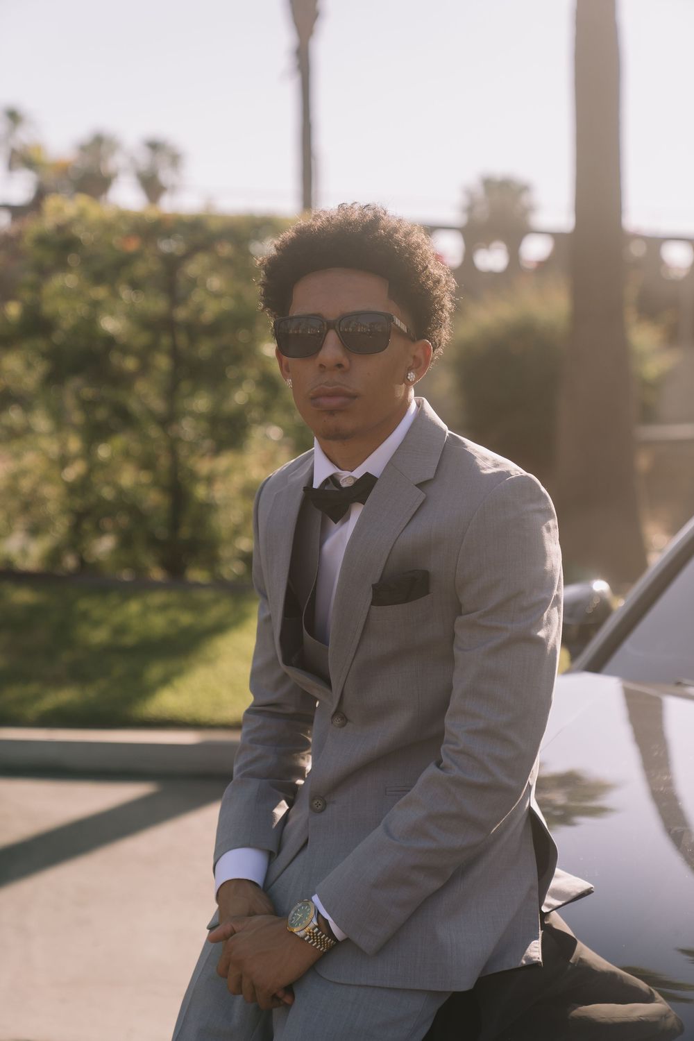 Young man in gray suit, bow tie, and sunglasses leaning on car outdoors.