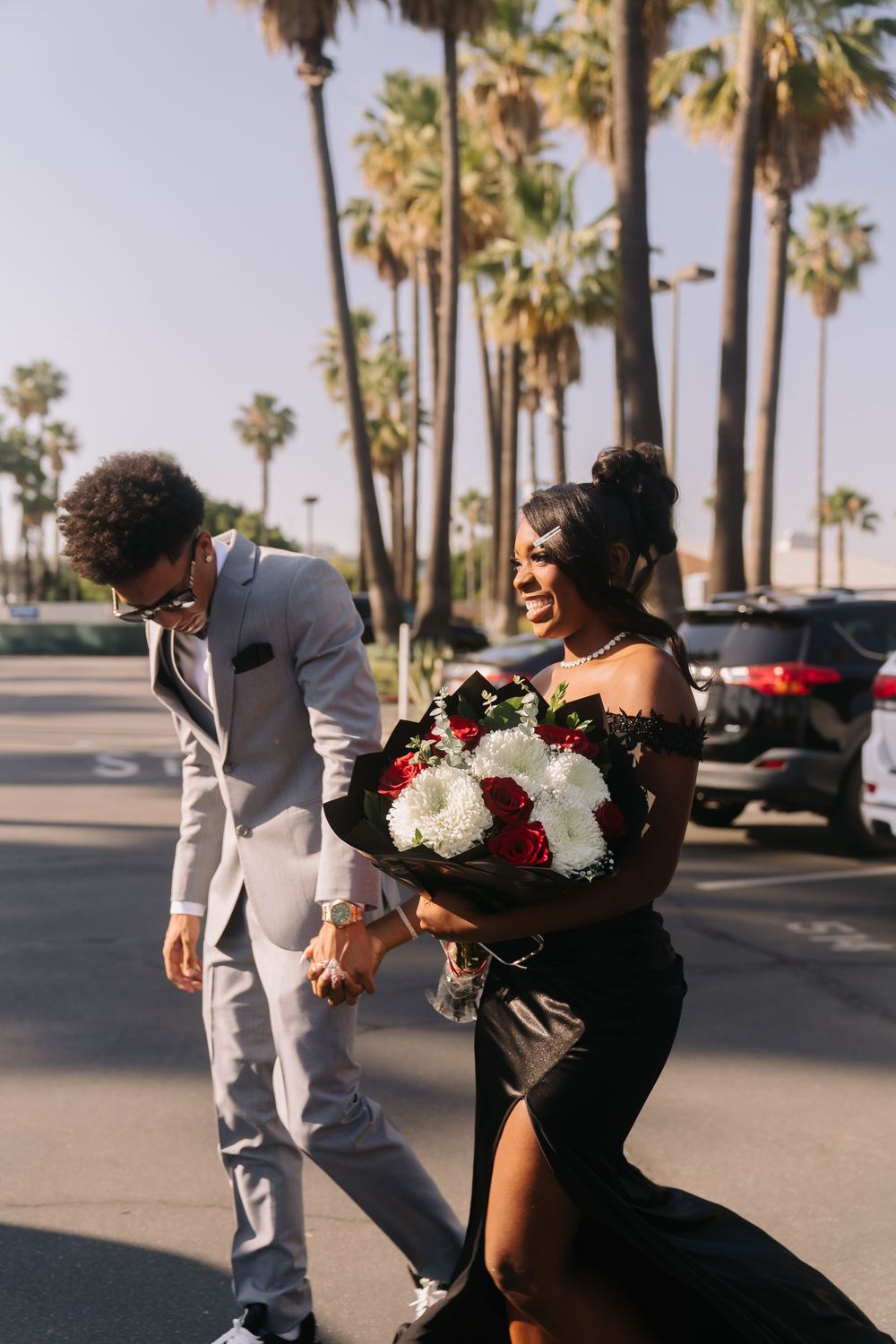 Couple in formal wear walking outdoors, woman holding red and white bouquet.