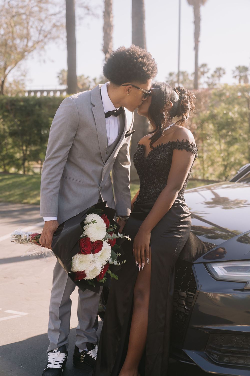 Couple in prom attire kissing by a car, man in gray suit with bouquet, woman in black gown, palm trees background.